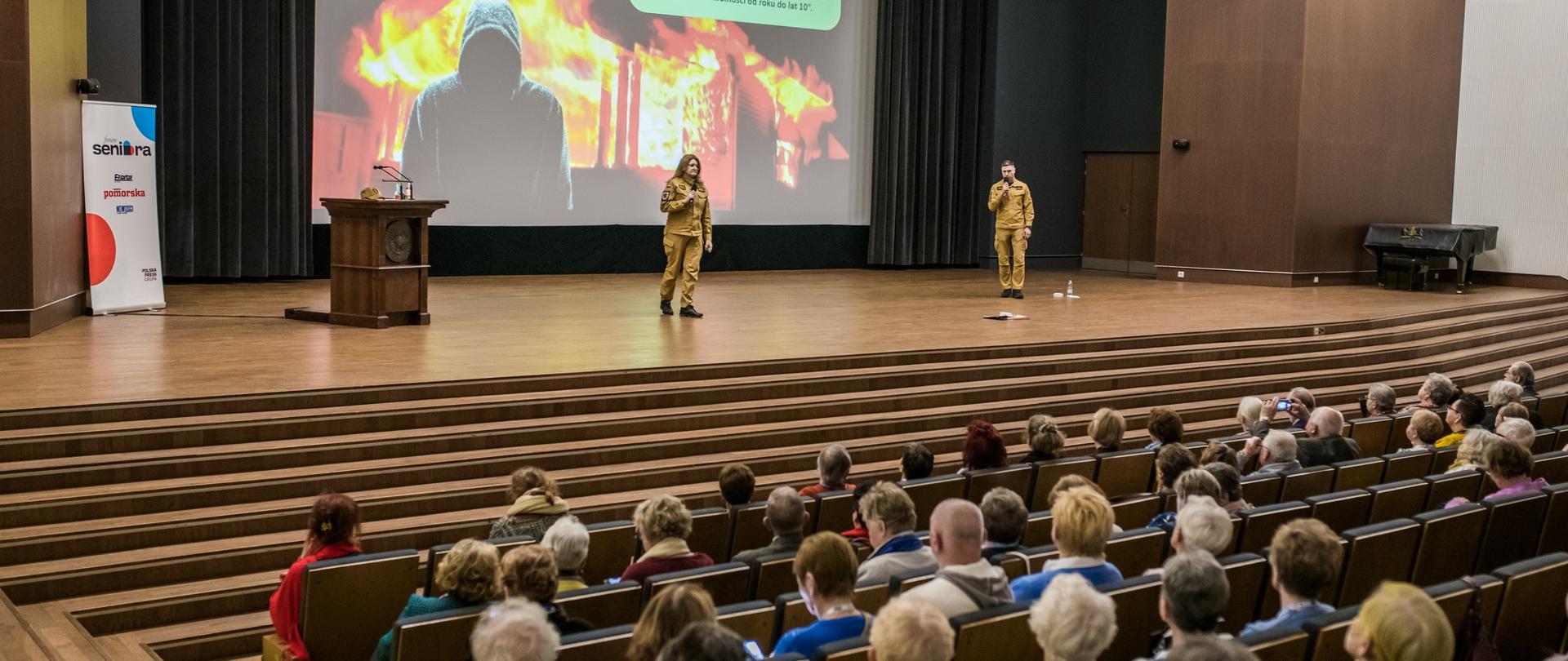 Na scenie Auli UMK stoi dwoje strażaków w mundurach służbowych. Za nimi wielki ekran, a na nim slajd z ogniem. Z przodu fragment widowni z uczestnikami wydarzenia.