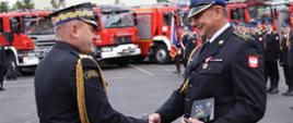 Zdjęcie zrobione na zewnątrz podczas uroczystości. Oficer Państwowej Straży Pożarnej w stopniu generała podaje rękę funkcjonariuszowi Państwowej Straży Pożarnej gratulując nagrody. Za nimi stoją ustawieni strażacy Państwowej Straży Pożarnej wraz z pocztem sztandarowym. W tle stoją ustawione samochody ratowniczo-gaśnicze. 