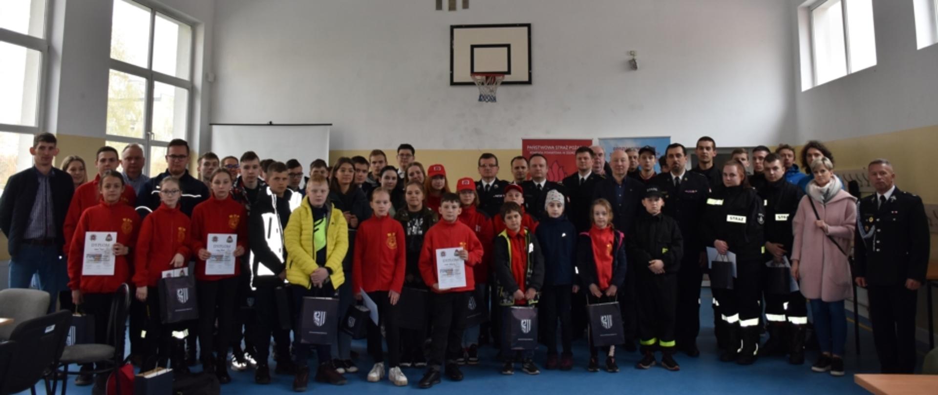 Dzień, Zdjęcie wykonane na tle ściany w sali gimnastycznej, na ścianie wisi tablica z koszem do gry w koszykówkę. Zdjęcie przedstawia grupę dzieci uczestniczących w OTWP, ich opiekunów, druhów OPS w ubraniach koszarowych lub mundurach wyjściowych oraz funkcjonariuszy PSP w umundurowaniu wyjściowym. Część dzieci ubrana w czerwone bluzach MDP. Cześć dzieci trzyma dyplomy.