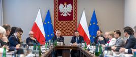 Kierownictwo MSWiA siedzi przy stole wspólnie z wojewodami. W tle duże polskie godło oraz flagi polskie i unijne 
