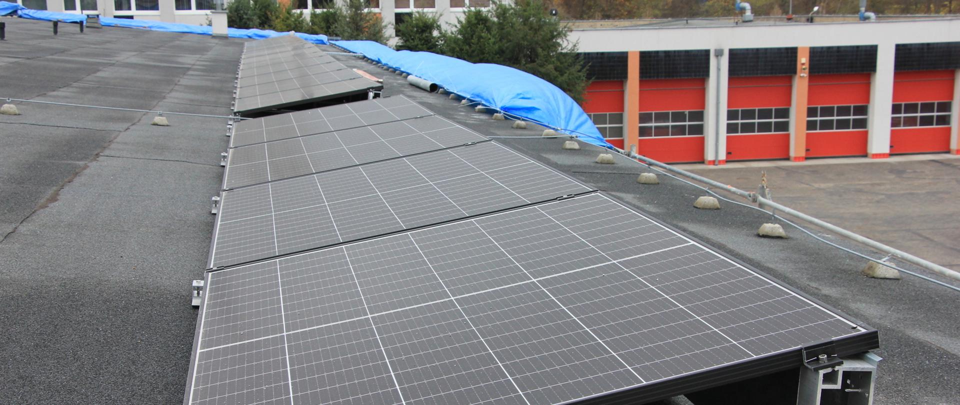 Panele fotowoltaiczne zainstalowane na dachu budynku. W tle widoczny budynek komendy.