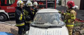 Na zdjęciu czterech strażaków stoi przy wraku pojazdu podczas szkolenia, w tle samochód pożarniczy