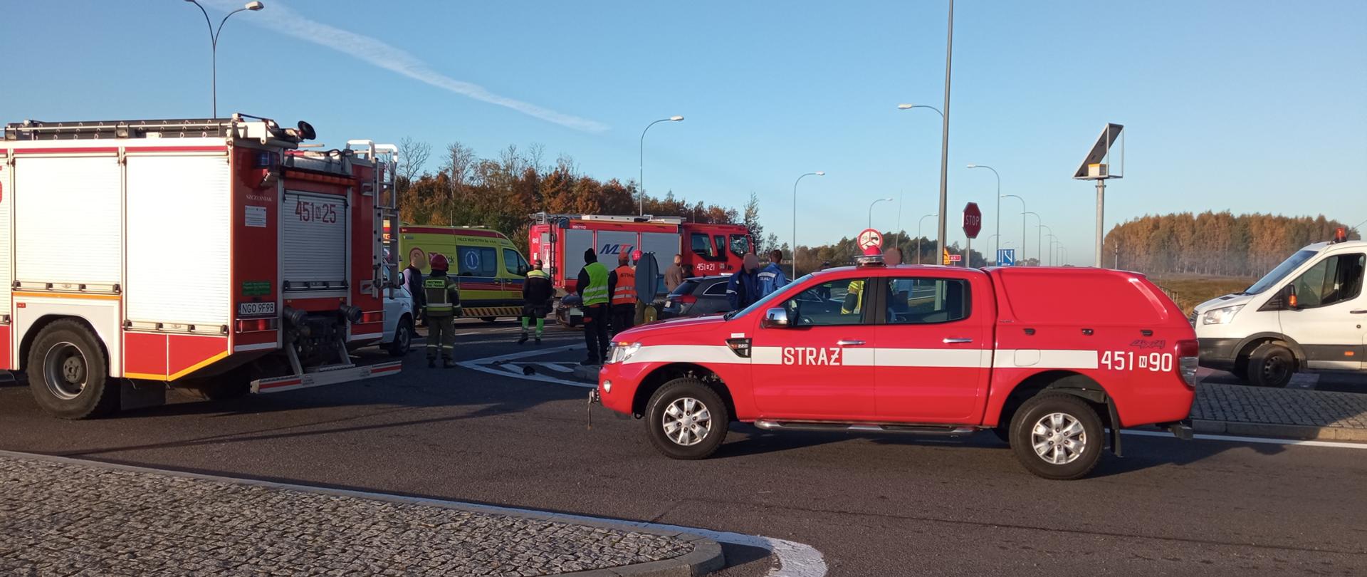 Zdjęcie przedstawia miejsce zderzenia samochodu osobowego i busa na obwodnicy Gołdapi. Obraz pokazuje trzy pojazdy pożarnicze, karetkę pogotowia oraz strażaków i osoby biorące udział w zdarzeniu
