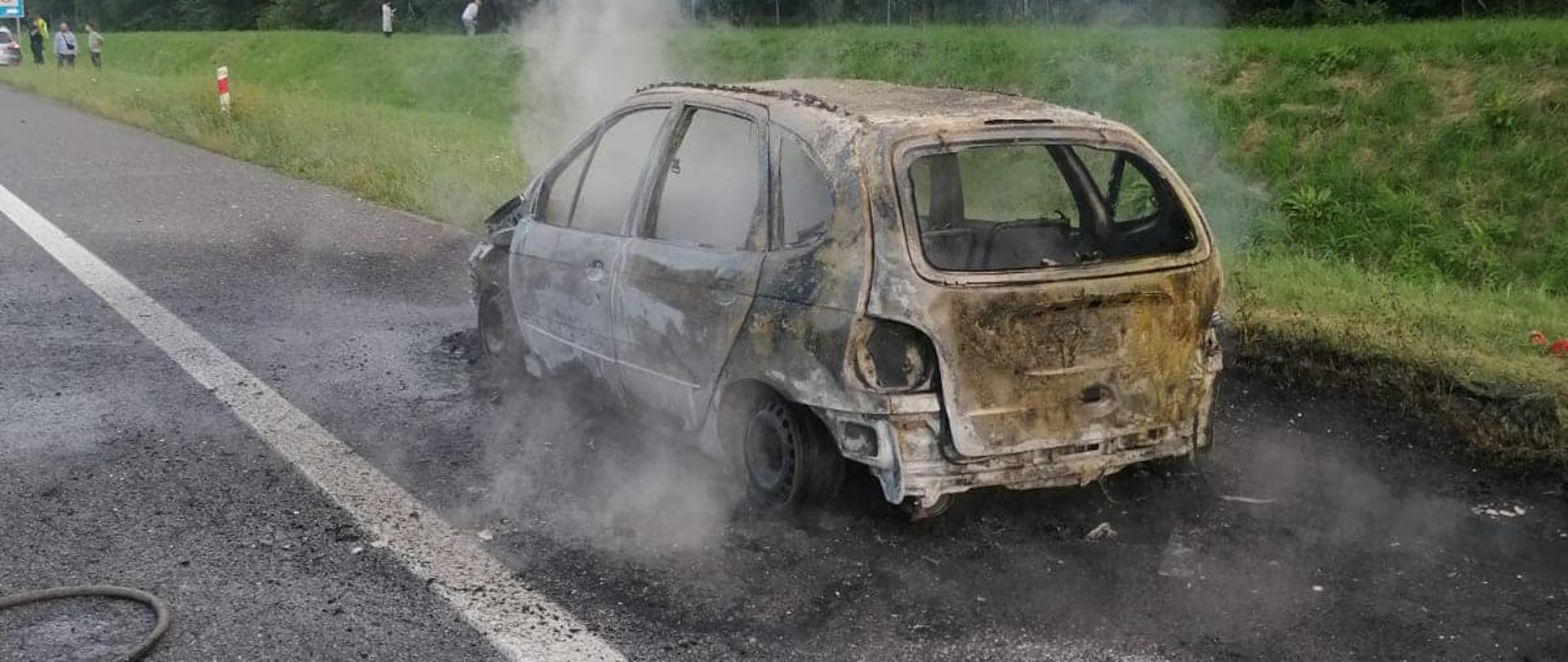 Na zdjęciu znajduje się wrak spalonego samochodu osobowego stojący na pasie awaryjnym autostrady