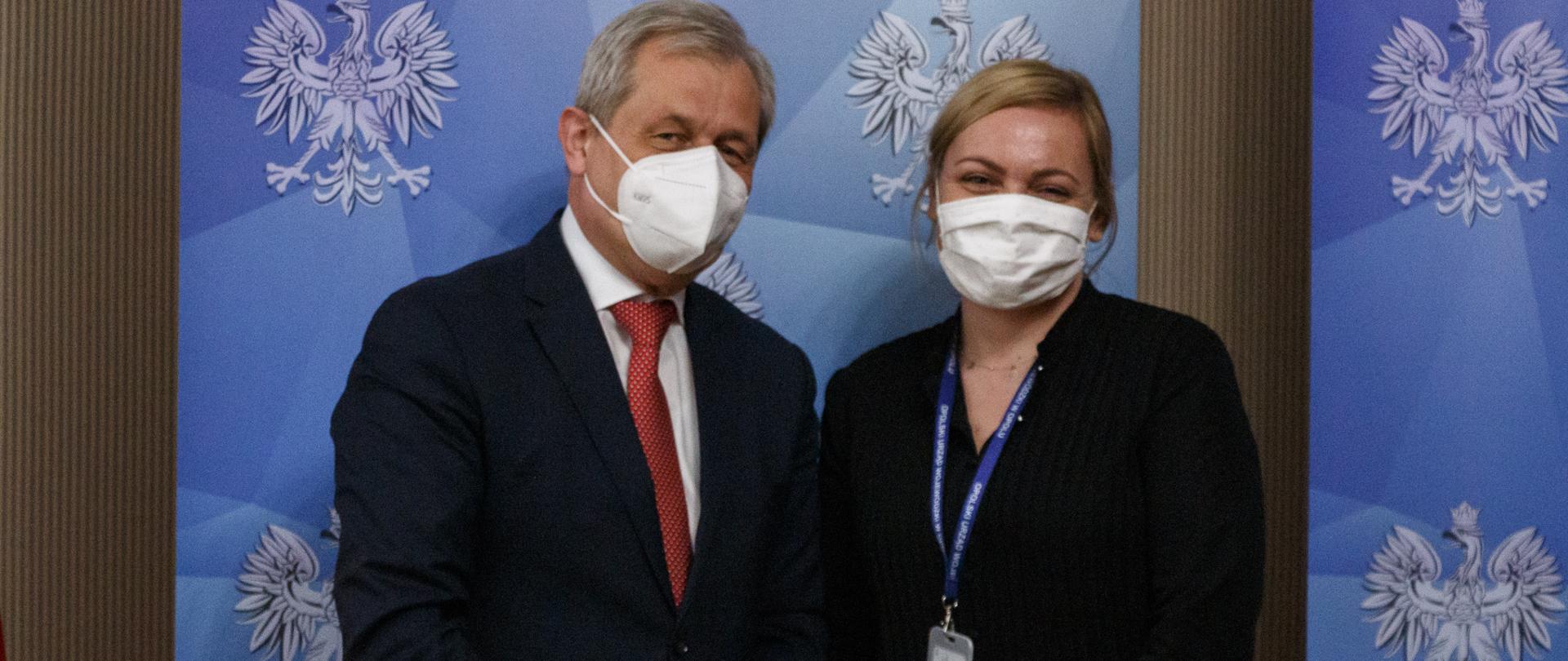 Na zdjęciu stoi wojewoda opolski ubrany w ciemny garnitur, a obok stoi kobieta ubrana w czarną sukienkę. Pozują do zdjęcia. Za nimi roll up Wojewoda Opolski w kolorze granatowym z godłem RP.