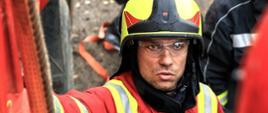 Na zdjęciu widać strażaka w umundurowaniu bojowym w hełmie oraz okularach. Hełm koloru żółtego. Strażak znajduje się obok wozu bojowego. Strażak jest instruktorem. Umundurowanie koloru czerwono-żółtego. 