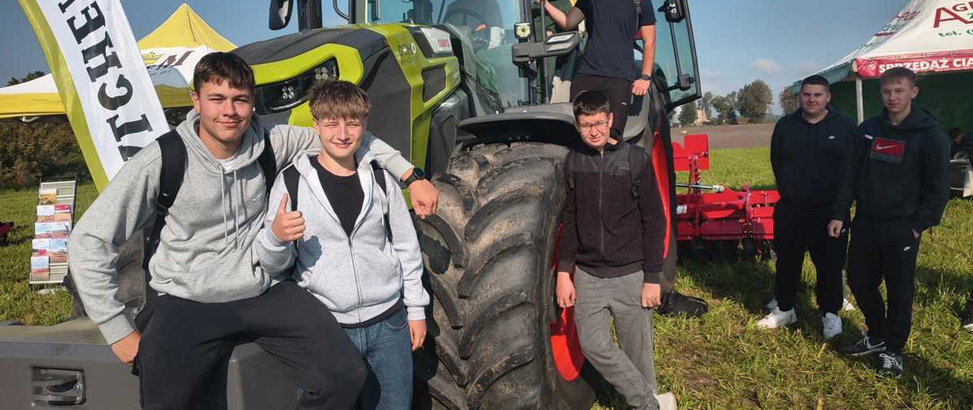 "Zdjęcie przedstawia grupę sześciu nastoletnich chłopców pozujących na tle dużego, nowoczesnego traktora rolniczego. Scena rozgrywa się na trawiastym polu w słoneczny dzień pod bezchmurnym, niebieskim niebem. Impreza ma charakter plenerowych targów rolniczych, na co wskazują widoczne w tle namioty reklamowe oraz flagi.
Oto szczegółowy opis rozmieszczenia osób i obiektów na zdjęciu:
• Centralny punkt (Traktor): Na środku kadru stoi potężny ciągnik rolniczy marki Claas (zielono-jasnoszary korpus z charakterystycznymi czerwonymi felgami). Uwagę zwraca jego ogromne, czarne koło z głębokim bieżnikiem, które znajduje się na pierwszym planie.
• Grupa po lewej stronie: Dwóch chłopców siedzi na przednim obciążniku traktora.
• Chłopak po lewej ma na sobie szarą bluzę z kapturem i czarne spodnie.
• Chłopak obok niego, również w szarej rozpinanej bluzie i szerokich jeansach, uśmiecha się i pokazuje kciuk w górę.
• Grupa na traktorze i obok koła:
• Trzeci nastolatek w ciemnej bluzie i szarych dresach stoi oparty bezpośrednio o wielką oponę ciągnika.
• Czwarty chłopak (w czarnym t-shircie) stoi wyżej, na stopniu prowadzącym do otwartej kabiny traktora, trzymając się stabilnie ramy pojazdu. W samej kabinie, za szybą, majaczy sylwetka kolejnej osoby.
• Grupa po prawej stronie: W tle, po prawej stronie maszyny, stoi dwóch kolejnych nastolatków ubranych na sportowo w ciemne bluzy i spodnie dresowe. Obaj patrzą w stronę obiektywu.
• Otoczenie: Po lewej stronie kadru widoczna jest biała flaga reklamowa z fragmentem czarnego napisu (końcówka „TCHEW”), a po prawej stronie w tle rozstawione są namioty wystawowe (w tym jeden biało-czerwony). Całość oświetla mocne, naturalne światło słoneczne."