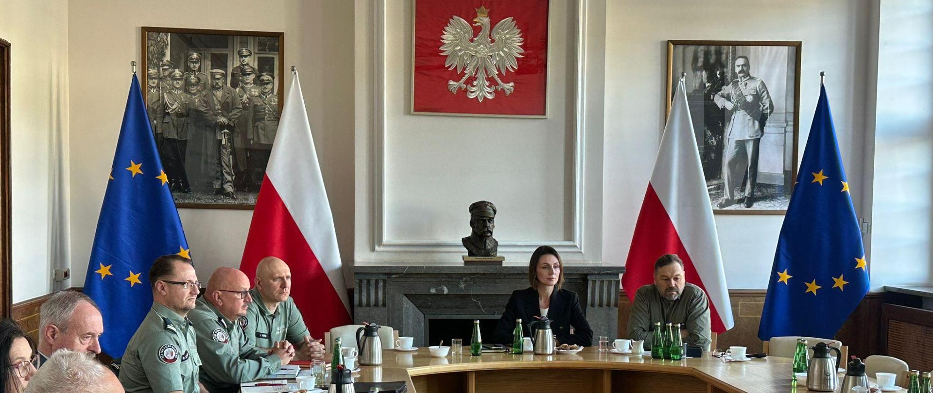 przy okrągłym stole siedzi kilka osób i dyskutuje, na ścianie godło Polski i obrazy przedstawiające marszałka Piłsudskiego