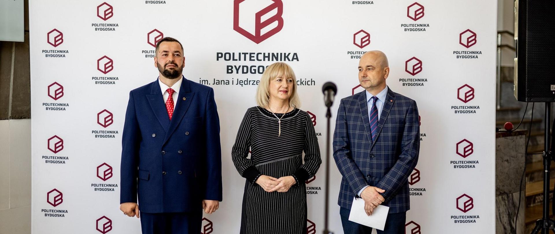 Pod ścianką z napisem Politechnika Bydgoska stoi kobieta w czarnej sukience pomiędzy dwoma mężczyznami w garniturach.