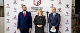 Pod ścianką z napisem Politechnika Bydgoska stoi kobieta w czarnej sukience pomiędzy dwoma mężczyznami w garniturach.