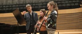 Prowadzący i dwie uczennice grające na klarnetach stoją przy pulpicie na scenie sali koncertowej PSM