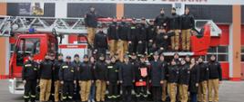 Strażacy w mundurach stoją w dwuszeregu na tle drabiny mechanicznej.
