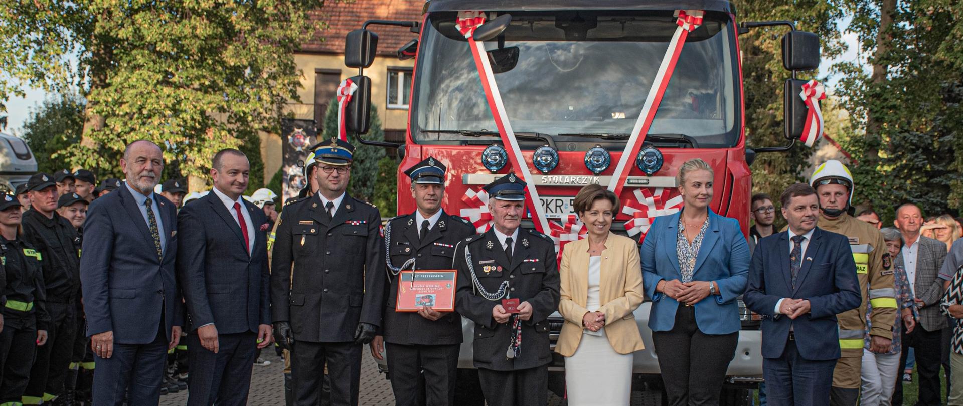 gruoa ludzi stoi przed samochodem strażackim, który ma na szybie wstęgę biało czerwoną