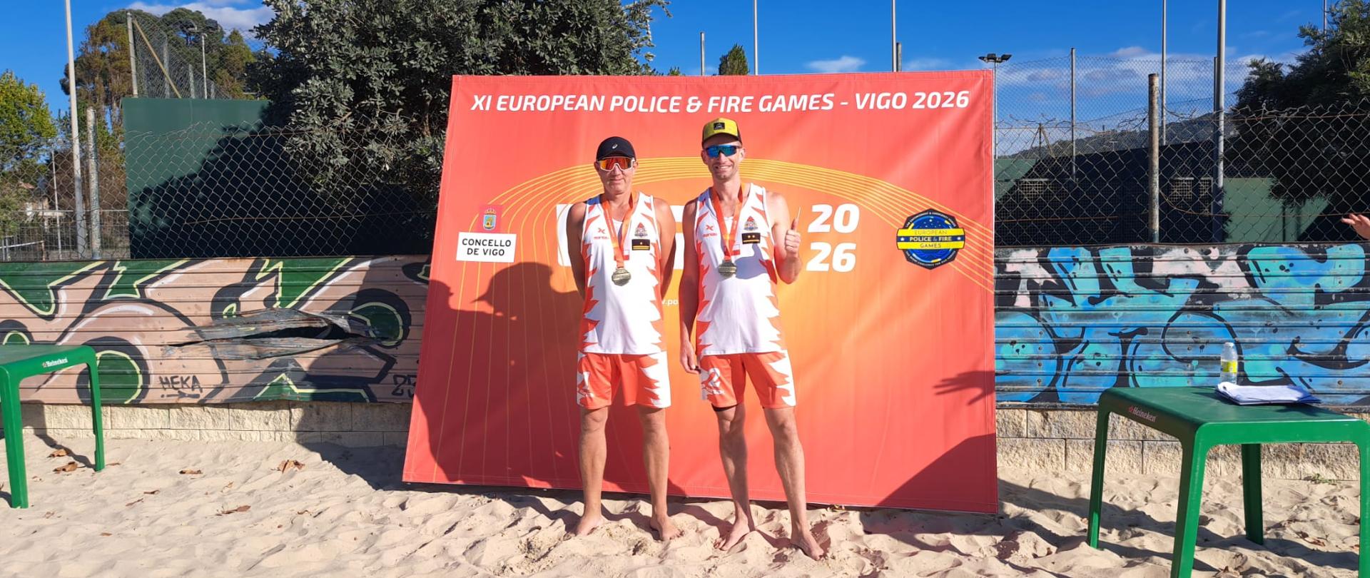 Dwaj polscy zawodnicy siatkówki plażowej stoją boso na piasku przed czerwoną ścianką promocyjną z napisami imprezy w Vigo. Obaj mają na sobie dopasowane stroje reprezentacji (biało-pomarańczowe koszulki i spodenki), okulary przeciwsłoneczne, czapeczki z daszkiem oraz złote medale zdobyte w turnieju. Jeden z nich pokazuje kciuk w górę.