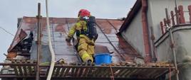 Na zdjęciu widać strażaka w aparacie powietrznym, który stoi na rusztowaniu postawionym przy ścianie budynku. Strażak ocenia stan dachu i sprawdza miejsce pożaru. Nad nim widać kosz autodrabiny w którym znajdują się strażacy z wężem pożarniczym gaszący pożar