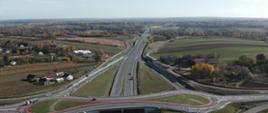 Widok z lotu ptaka na węzeł drogowy typu bezkolizyjnego: dwujezdniowa droga z pasem rozdziału, wiaduktem nad drogą główną, rondami turbinowymi oraz łącznicami wjazdowymi i zjazdowymi; otoczenie stanowią tereny rolnicze i rozproszona zabudowa