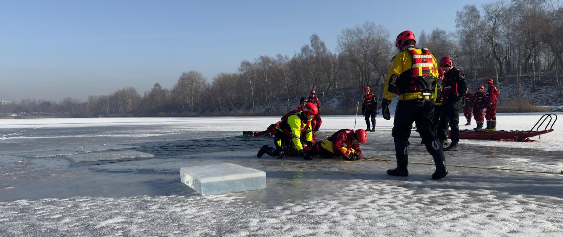 Na zamarzniętym jeziorze strażacy w czerwonych i żółtych suchych skafandrach i kaskach strojach ratowniczych ćwiczą akcję lodową. Kilku z nich klęczy przy przeręblu, wyciągając osobę z wody na lód i zabezpieczając ją liną. Inni stoją w pobliżu i obserwują przebieg ćwiczeń. Na lodzie widać wycięty blok lodu i sanie ratownicze, a w tle znajdują się nagie, zimowe drzewa i jasne niebo.