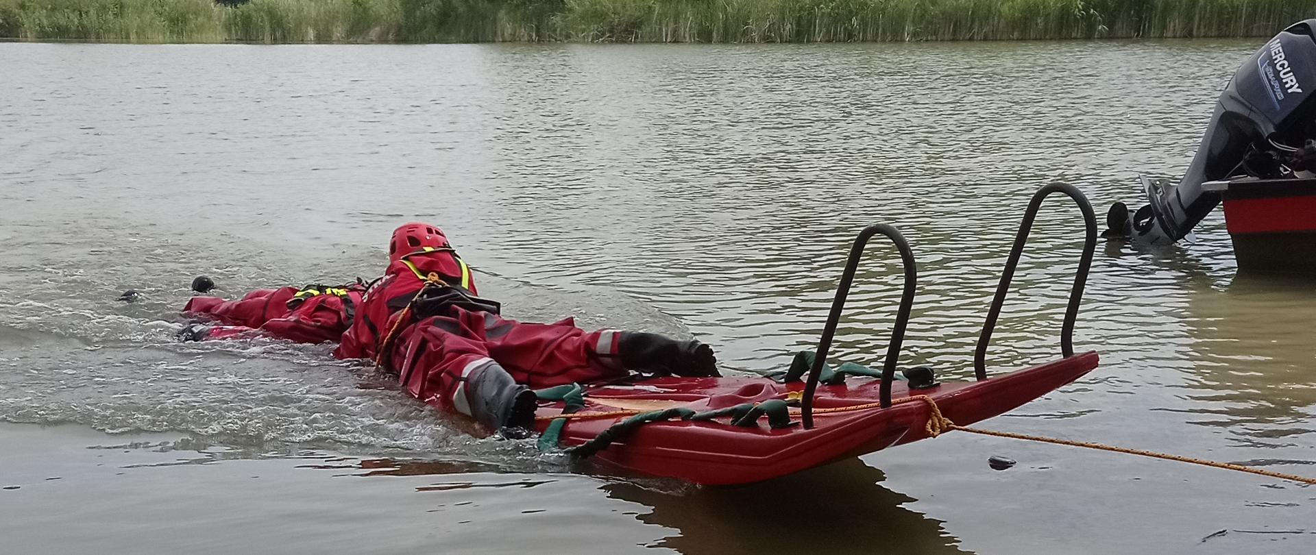 Zdjęcie wykonane z brzegu, na którym widać ratowników ubranych w czerwone skafandry wypornościowe, czarne buty i czerwone kaski. Ratownicy znajdują się na pływających saniach lodowych wykonanych z tworzywa. Po prawej stronie fotografii stoi zielono-czerwona łódź ratownicza z silnikiem zaburtowym. W tle widać wysokie drzewa oraz roślinność przy brzegową.