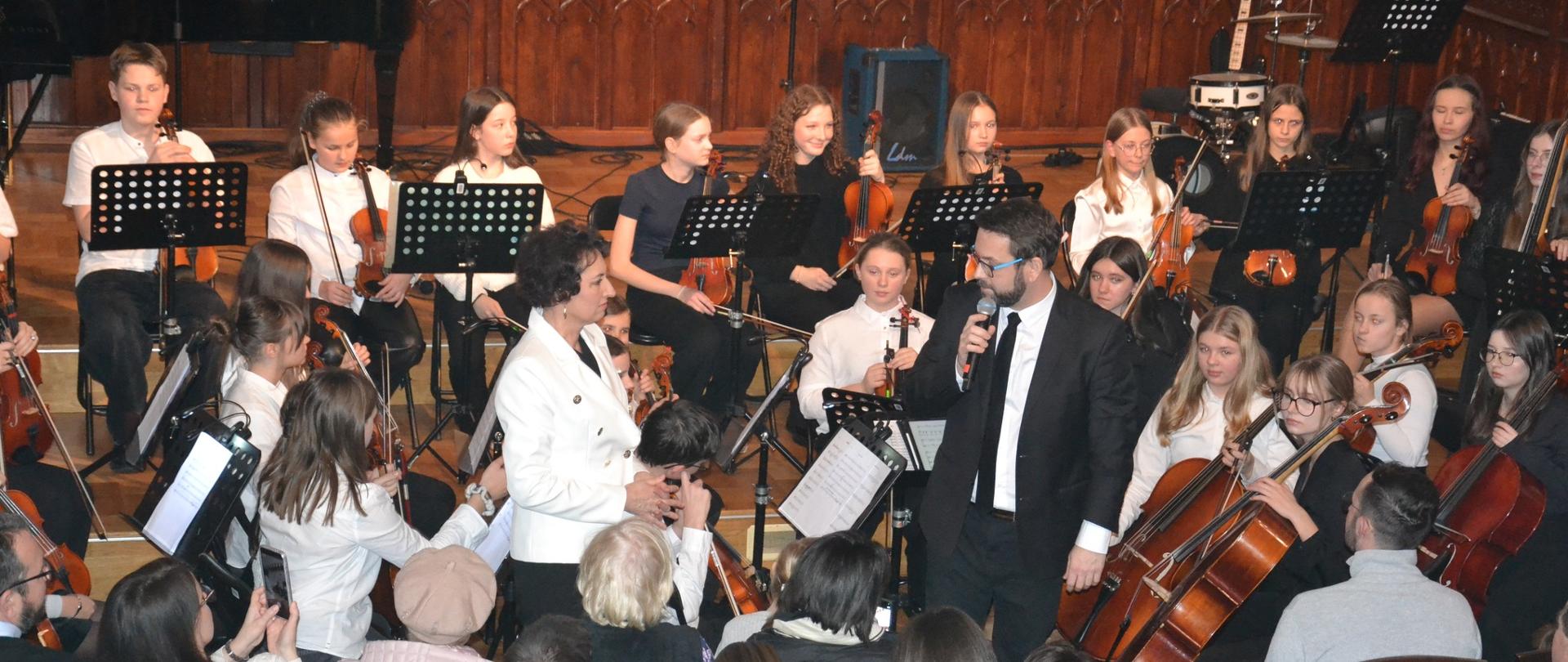 Fotografia przedstawia finał koncertu w Sali Królewskiej – na scenie występuje orkiestra smyczkowa dzieci i młodzieży z dyrygentem oraz prowadzącym koncert z mikrofonem. Na pierwszym planie widoczna jest publiczność, a całość oddaje uroczystą atmosferę wspólnego muzykowania.