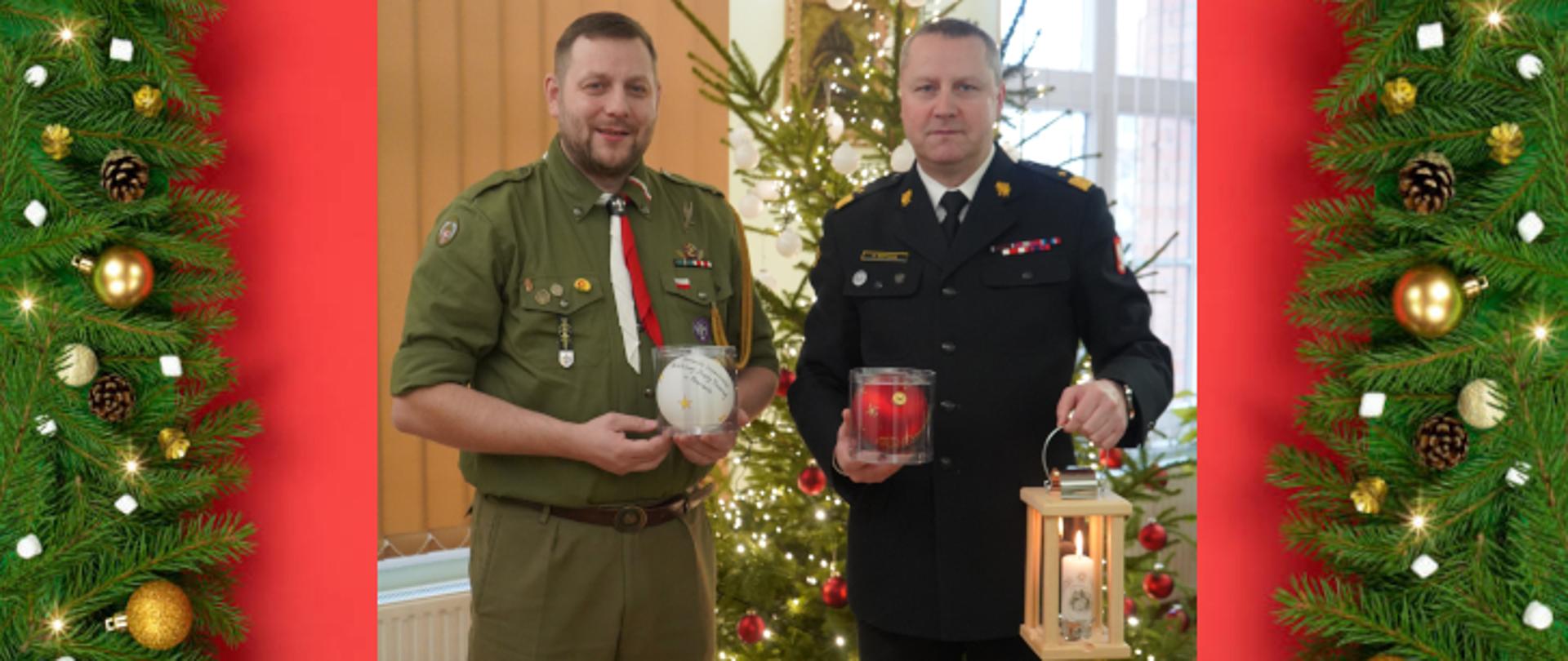 mężczyzna harcerz w zielonym mundurze trzyma białą duża bombkę, obok stoi strażak w ciemnym mundurze w ręku trzyma lampion z zapaloną świeczką a w drugiej czerwoną bombkę , w tle choinka z biało czerwonymi lampkami
