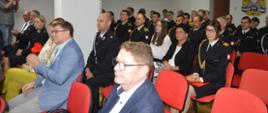 Świetlica komendy. Na krzesłach siedzą uczestnicy zbiórki, osoby cywilne oraz strażacy w mundurach wyjściowych. 