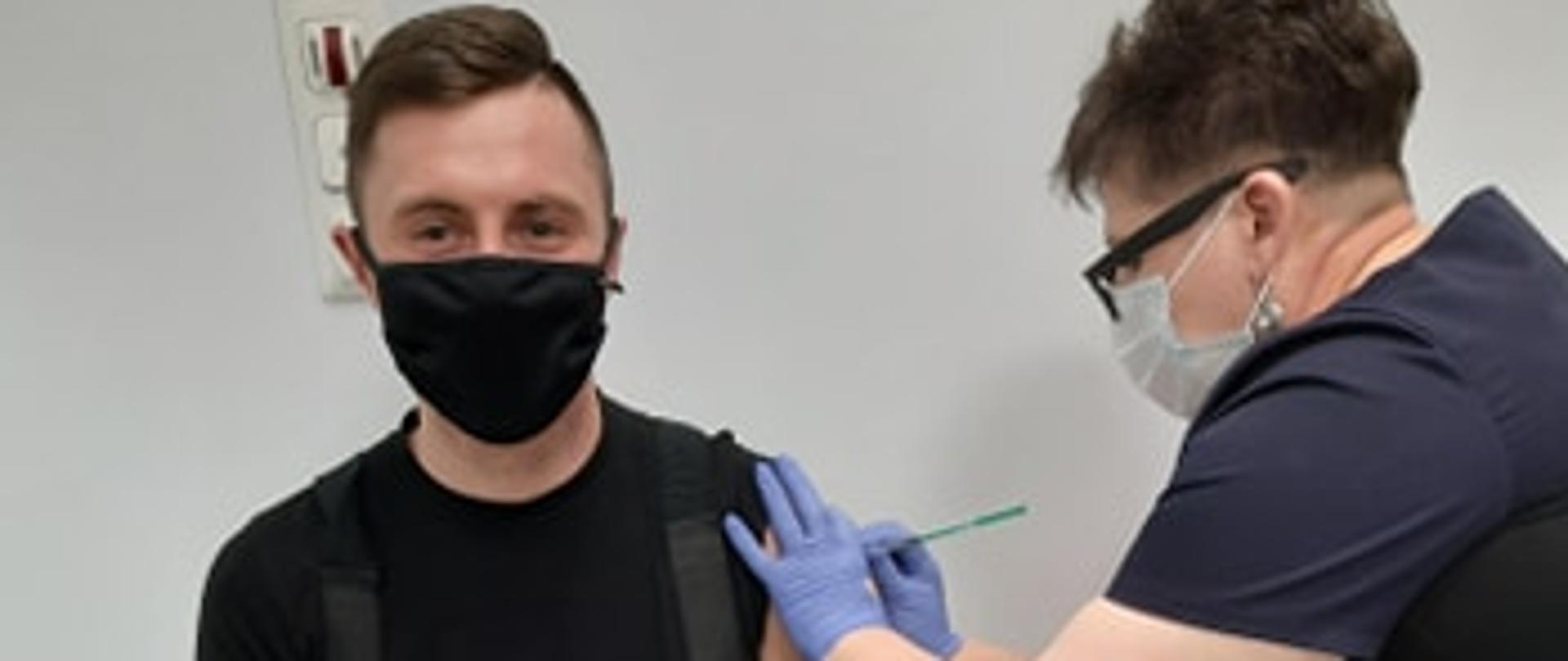 Zdjęcie przedstawia strażaka poddającego się szczepieniu przez naczelną pielęgniarką Szpitala w Opatowie. Strażak i pielęgniarka w maseczkach ochronnych oraz w rękawiczkach nitrylowych.