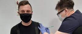 Zdjęcie przedstawia strażaka poddającego się szczepieniu przez naczelną pielęgniarką Szpitala w Opatowie. Strażak i pielęgniarka w maseczkach ochronnych oraz w rękawiczkach nitrylowych.