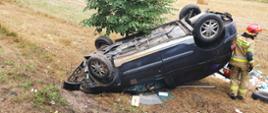 Na poboczu drogi dachował samochód marki Renault Scenic. Samochodem podróżowały dwie osoby. Strażak zabezpiecza teren działań.