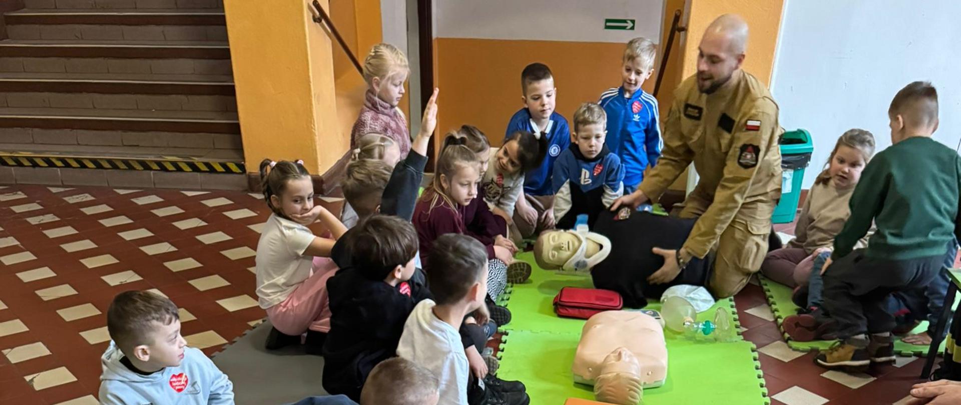 Na zdjęciu widoczne są dzieci, które ćwiczą udzielanie pierwszej pomocy na fantomie. Zajęcia prowadzi strażak.