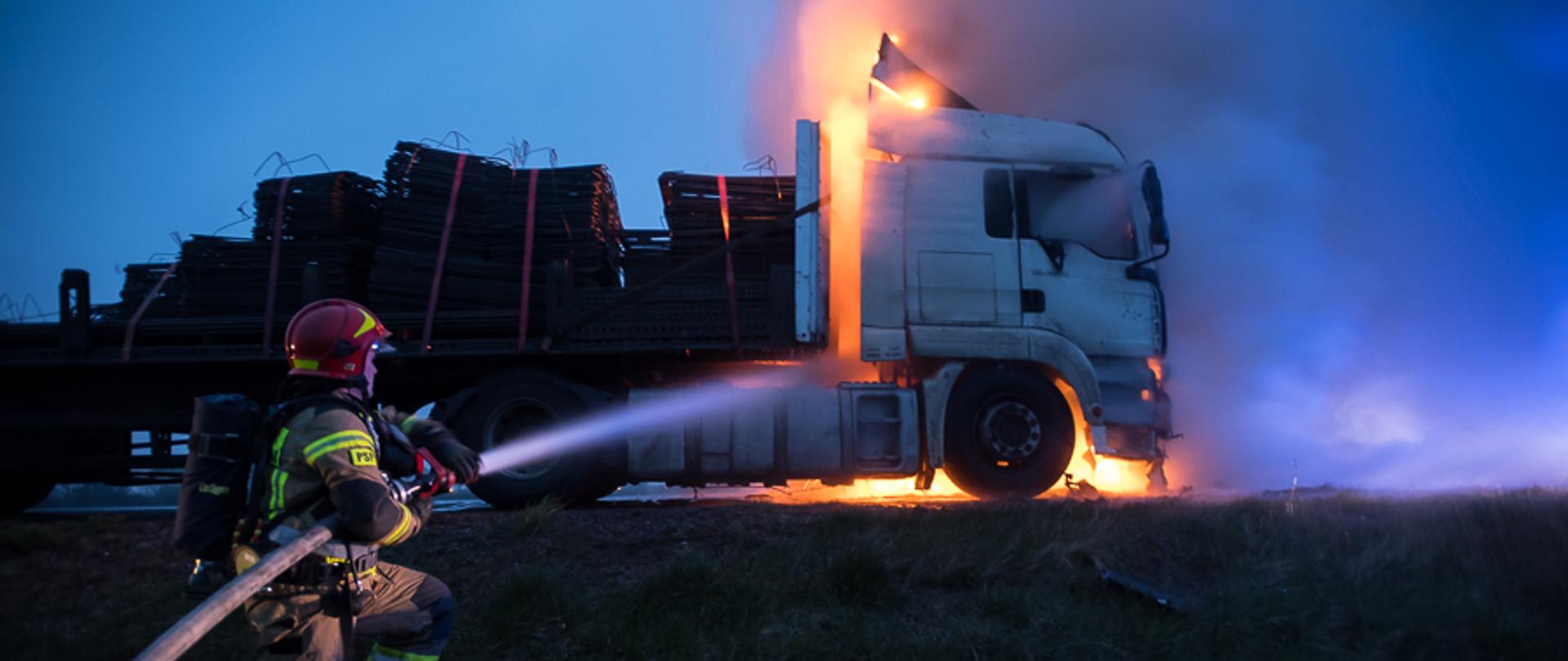 Na zdjęciu widoczny strażak PSP w piaskowym ubraniu specjalnym i czerwonym hełmie, z aparatem powietrznym na plecach. W rękach trzyma linię wężową i podaje wodę na palącą się kabinę ciężarówki.
