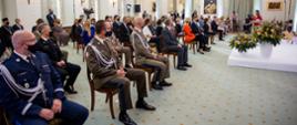 Na zdjęciu: Prezydent RP wraz z Pierwszą Damą, wysocy rangą oficerowie różnych służb. Siedzą na krzesłach. Są w maseczkach. Na scenie przemawia młoda kobieta. Obok sceny duże bukiety kwiatów.. 