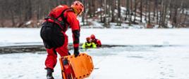 Strażak z deską ortopedyczną idzie po tafli lodu do przerębla w którym są strażacy pozorujący osoby poszkodowane