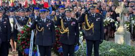 Trzech strażaków Państwowej Straży Pożarnej oddaje honor za nimi stoją osoby funkcjonariusze służb podległych Ministerstwu Spraw Wewnętrznych i Administracji oraz osoby biorące udział w uroczystościach pogrzebowych. 