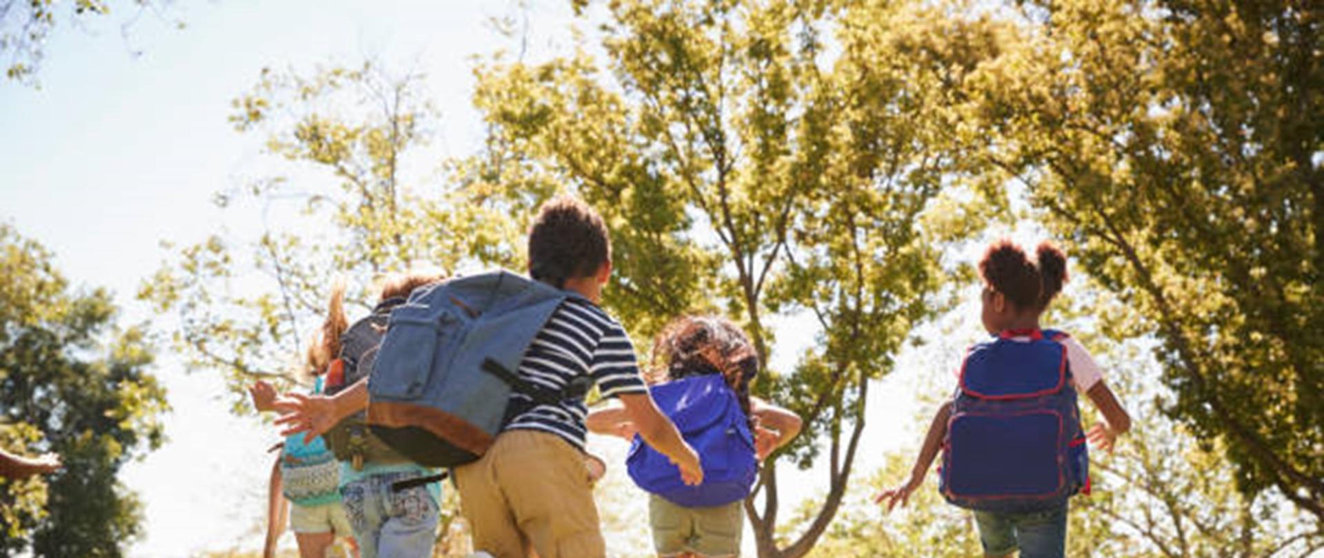 Dzieci z plecakami biegnące po trawie, wśród drzew.