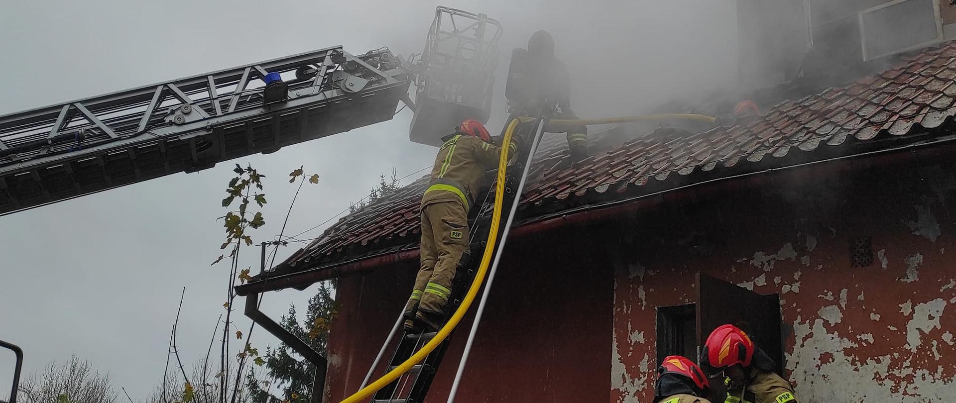 Na zdjęciu strażacy pracujący przy pożarze budynku mieszkalnego. Drabina mechaniczna i przystawna przy dachu budynku. Strażacy z pomieszczenia wynoszą rzeczy. Nad budynkiem unosi się biały dym.