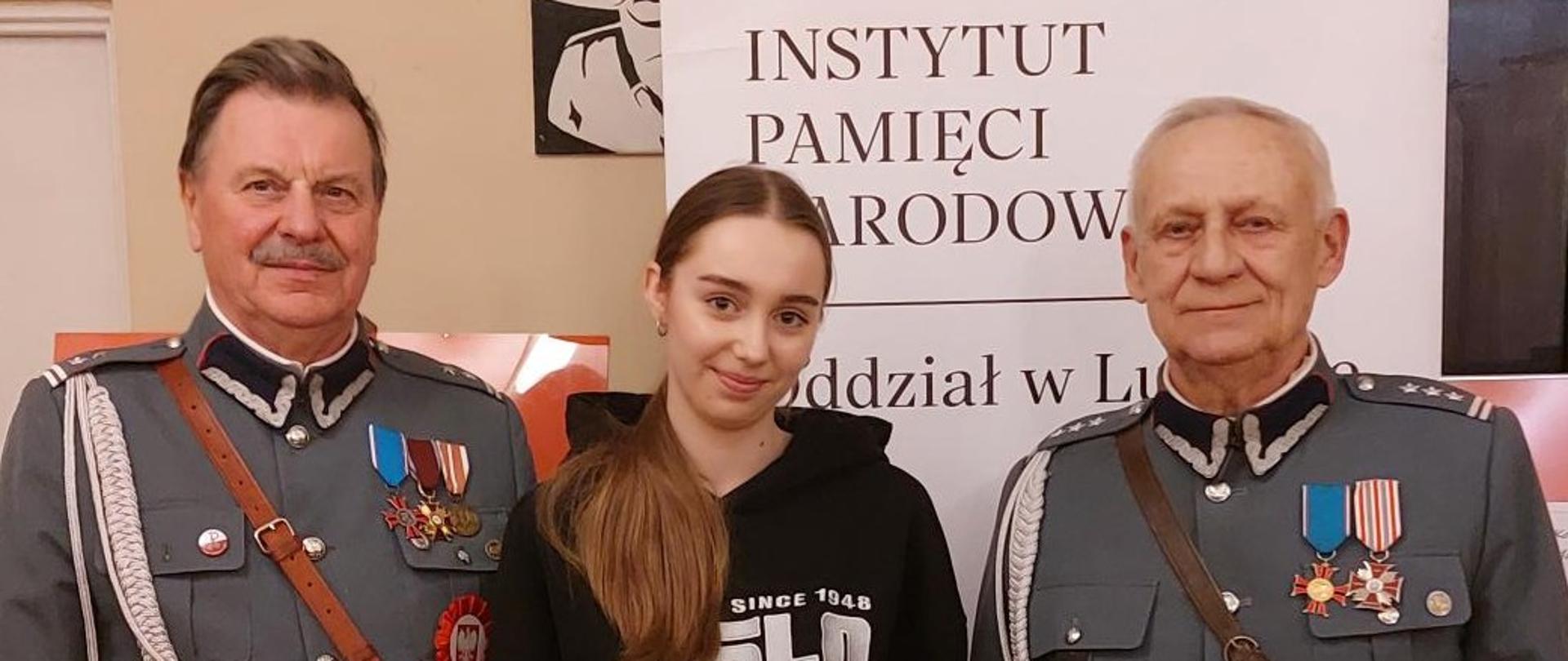 Na środku stoi dziewczyna trzymająca w ręku "DYPLOM dla Zuzanny Lis za zajęcie I miejsca" oraz czarną torbę. Obok niej - po prawej i po lewej stronie stoją starsi mężczyźni w szarych wojskowych mundurach z medalami na piersiach. W tle widać fragmenty białego baneru z czarnym napisem INSTYTUT PAMIĘCI NARODOWEJ Oddział w Lublinie.