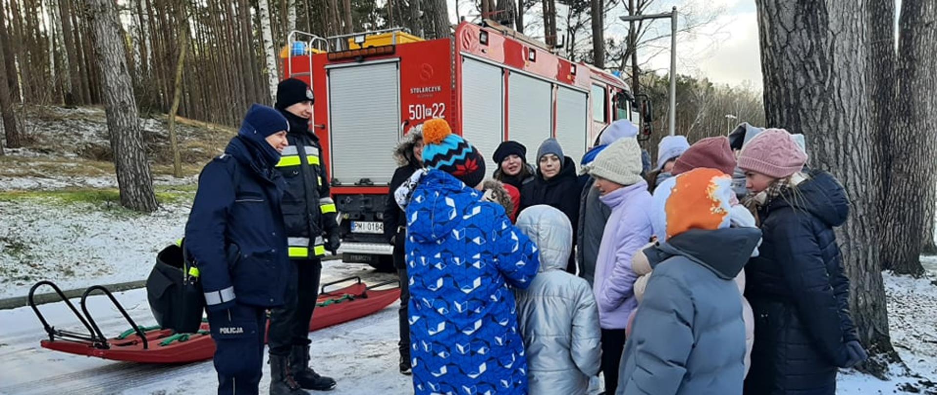 Zdjęcie przedstawia grupę dzieci i młodzieży, przy nich strażak i policjantka, którzy rozmawiają ze sobą. Obok samochód pożarniczy i sanie lodowe.