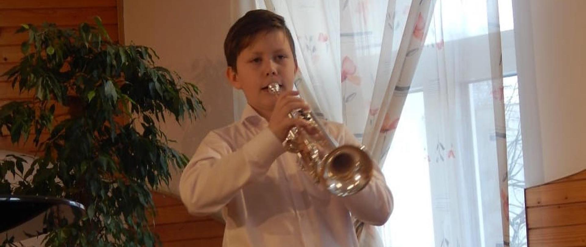 Chłopiec gra na trąbce, ubrany w białą koszulę i czarne spodnie. Stoi w auli szkoły muzycznej, w tle widać fortepian