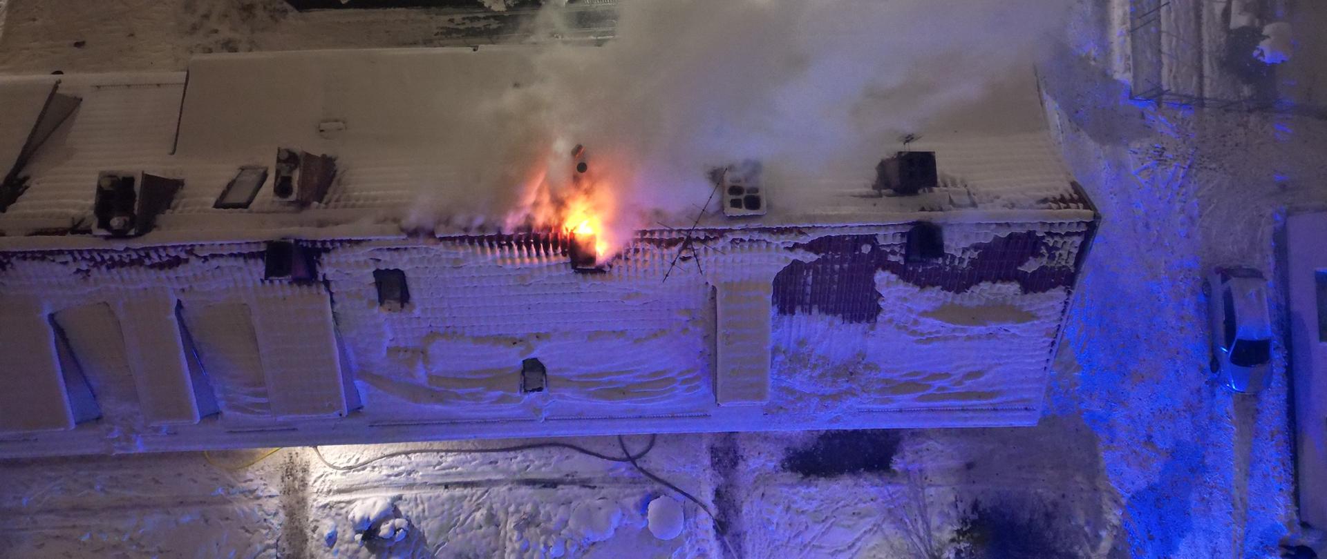 Widok z góry na dach wielorodzinnego budynku mieszkalnego. Dach i otoczenie dookoła jest zaśnieżone. Obok jednego z kominów wydostają się płomienie z palącego się poddasza. 