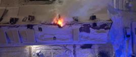 Widok z góry na dach wielorodzinnego budynku mieszkalnego. Dach i otoczenie dookoła jest zaśnieżone. Obok jednego z kominów wydostają się płomienie z palącego się poddasza. 