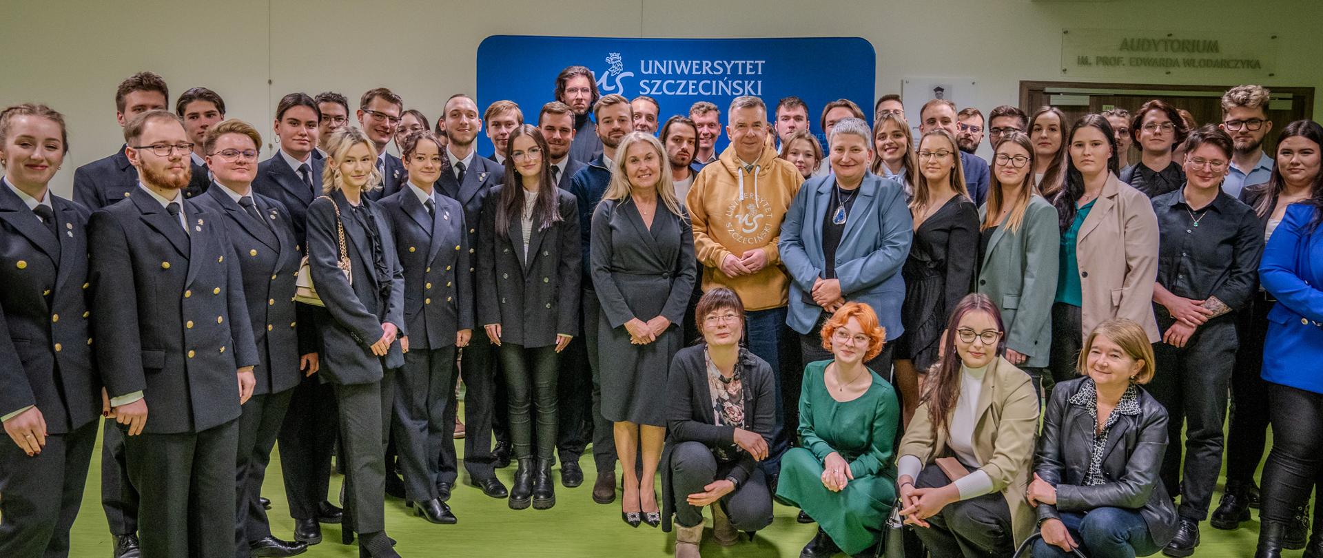 Pod niebieską ścianką z napisem Uniwersytet Szczeciński stoi duża grupa ludzi, pośrodku minister Wieczorek w żółtej bluzie z kapturem.