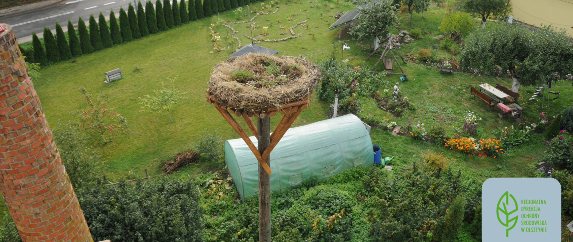 W centralnej części ogród z platformą, na której umiejscowione jest gniazdo bociana. Poniżej platformy szklarnia, obok której rosną kwiaty i drzewka, stoi huśtawka oraz stół z ławkami. W prawym dolnym rogu logo Regionalnej Dyrekcji Ochrony Środowiska w Olsztynie 