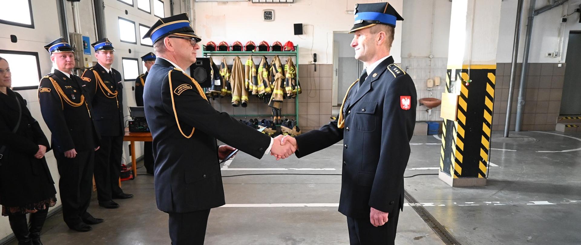 Na zdjęciu widać Komendanta Powiatowego PSP w Tucholi st. bryg. Dariusza Goźlińskiego wręczającego Decyzję o zwolnieniu ze służby asp. sztab. Wiesławowi Anhaltowi. Funkcjonariusze stoją naprzeciw siebie, bokiem do obiektywu aparatu w mundurach wyjściowych. Zdjęcie zrobione w garażu KP PSP w Tucholi.
