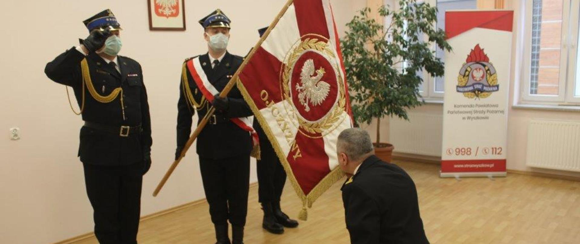 Komendant Powiatowy PSP Wyszków klęczy przed pochylonym sztandarem komendy całując go