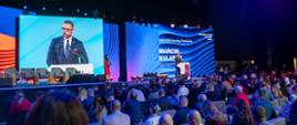 Zdjęcie z boku pod kątem, na podwyższeniu przed wielkim ekranem z napisem na niebieskim tle KNM 2026 Opening Ceremony - Marcin Kulasek stoi minister Kulasek i mówi do mikrofonu na stojaku, z boku stoi kobieta w czerwonej sukni, przed nimi wypełniona ludźmi sala.
