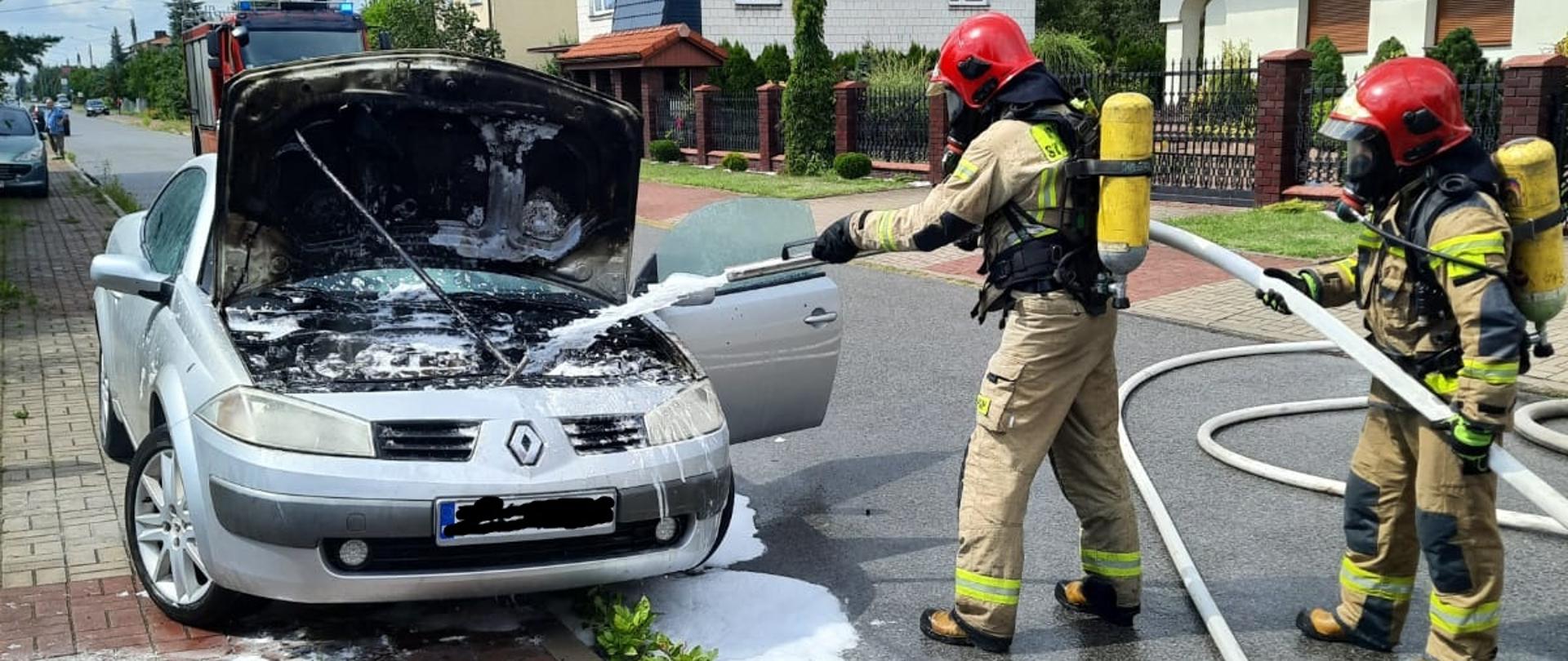 Zdjęcie obrazuje pożar komory silnika samochodu osobowego Renault. Dwóch strażaków ubranych w sprzęt ochrony układu oddechowego podaje prąd piany ciężkiej do silnika celem jego ugaszenia. Przed samochodem na chodniku leży wąż ogrodowy którym próbował gasić pożar właściciel. W oddali widać samochód pożarniczy.