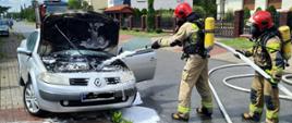 Zdjęcie obrazuje pożar komory silnika samochodu osobowego Renault. Dwóch strażaków ubranych w sprzęt ochrony układu oddechowego podaje prąd piany ciężkiej do silnika celem jego ugaszenia. Przed samochodem na chodniku leży wąż ogrodowy którym próbował gasić pożar właściciel. W oddali widać samochód pożarniczy.