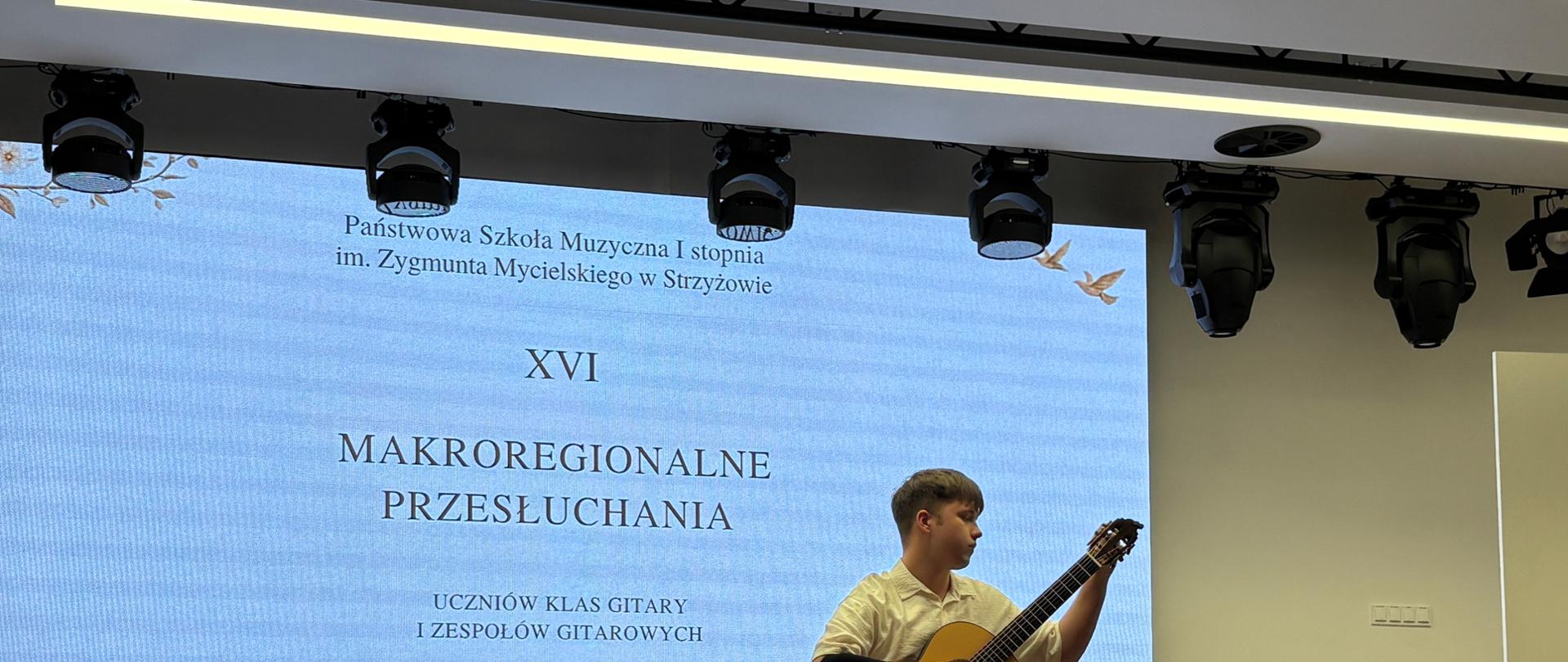 Na zdjęciu widać salę koncertową podczas XVI Makroregionalnych Przesłuchań Uczniów Klas Gitary i Zespołów Gitarowych w Strzyżowie. Na środku sceny siedzi Jan Szabat gitarzysta grający na gitarze klasycznej. Muzyk prezentuje utwór w skupieniu, przyjmując pozycję wykonawczą z podnóżkiem pod nogą.W tle znajduje się duży ekran, na którym wyświetlono nazwę wydarzenia, miejsce oraz datę: 28 maja 2026 roku. Nad sceną widoczny jest profesjonalny system oświetlenia scenicznego. Obok wykonawcy stoją krzesła i pulpit na nuty. Zdjęcie ukazuje moment występu konkursowego Jana Szabata podczas prezentacji programu przed jury i publicznością. 