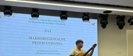 Na zdjęciu widać salę koncertową podczas XVI Makroregionalnych Przesłuchań Uczniów Klas Gitary i Zespołów Gitarowych w Strzyżowie. Na środku sceny siedzi Jan Szabat gitarzysta grający na gitarze klasycznej. Muzyk prezentuje utwór w skupieniu, przyjmując pozycję wykonawczą z podnóżkiem pod nogą.W tle znajduje się duży ekran, na którym wyświetlono nazwę wydarzenia, miejsce oraz datę: 28 maja 2026 roku. Nad sceną widoczny jest profesjonalny system oświetlenia scenicznego. Obok wykonawcy stoją krzesła i pulpit na nuty. Zdjęcie ukazuje moment występu konkursowego Szypulskiego podczas prezentacji programu przed jury i publicznością. 