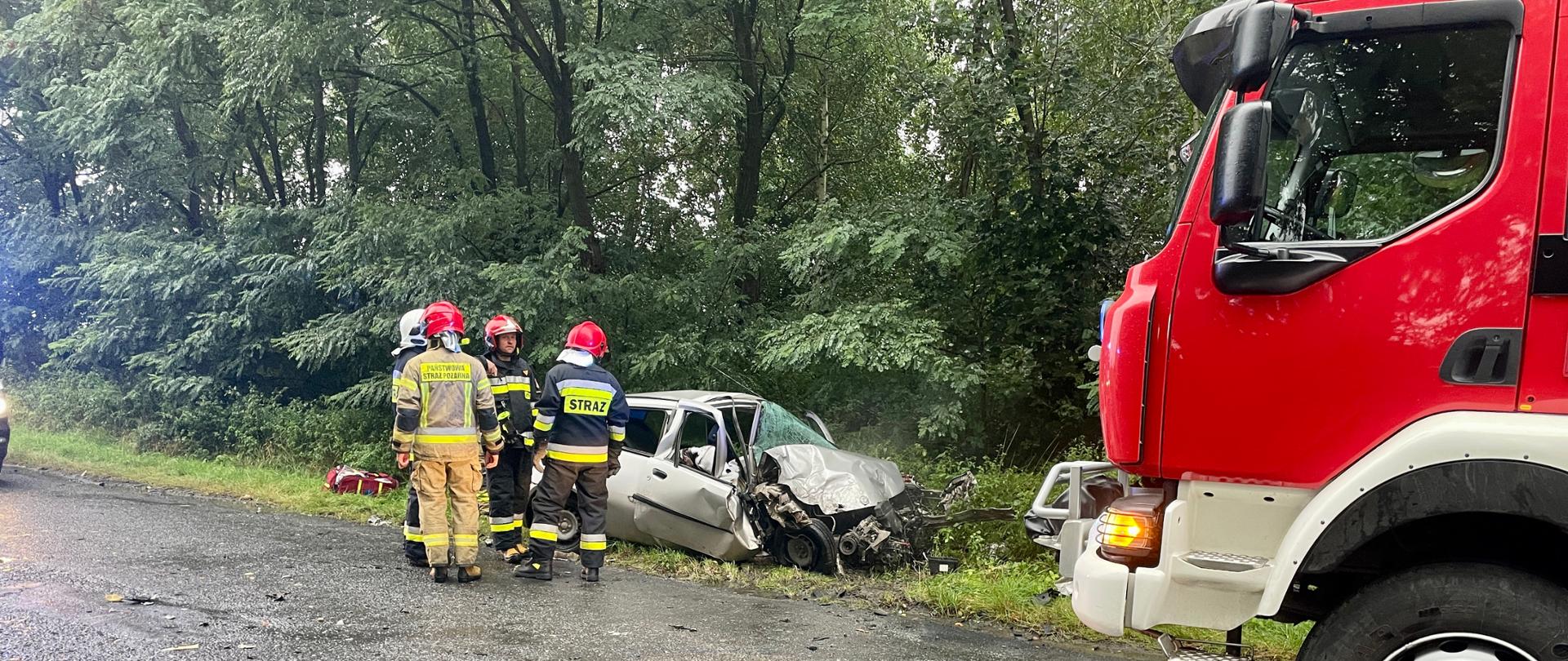strażacy stojący na tle wraku pojazdu osobowego, po prawej przód strażackiego wozu bojowego